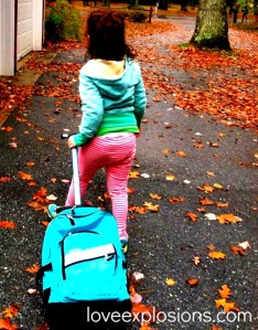 a school age child wearing a green hoody and pink and white striped pants pulls an aqua colored backpack on wheels up a driveway covered in leaves.