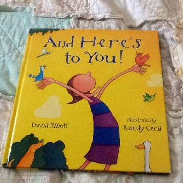 image is a yellow children's book.  There is an illustrated person with arms wide open.  The title of the book reads, " "And here's to you.